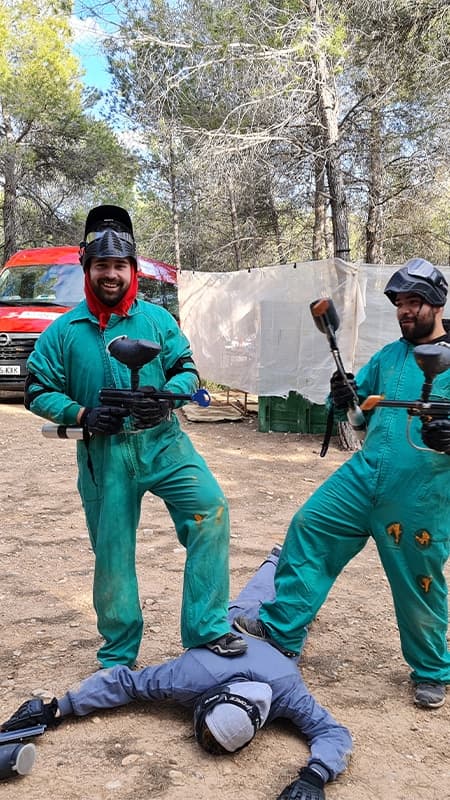 Jugador de paintball protegido con máscara y mono apuntando con su marcadora en un escenario exterior con obstáculos.